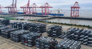 Rows of new cars stacked on racks at a Chinese port, with container cranes behind