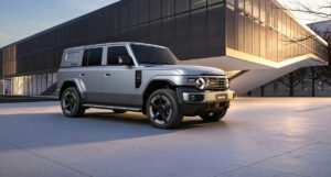 Silver iCAUR V25 boxy SUV, front three-quarter view, parked in front of a modern glass building at dusk