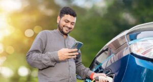 A man plugs a charging cable into an electric car at an outdoor charging station while using a smartphone